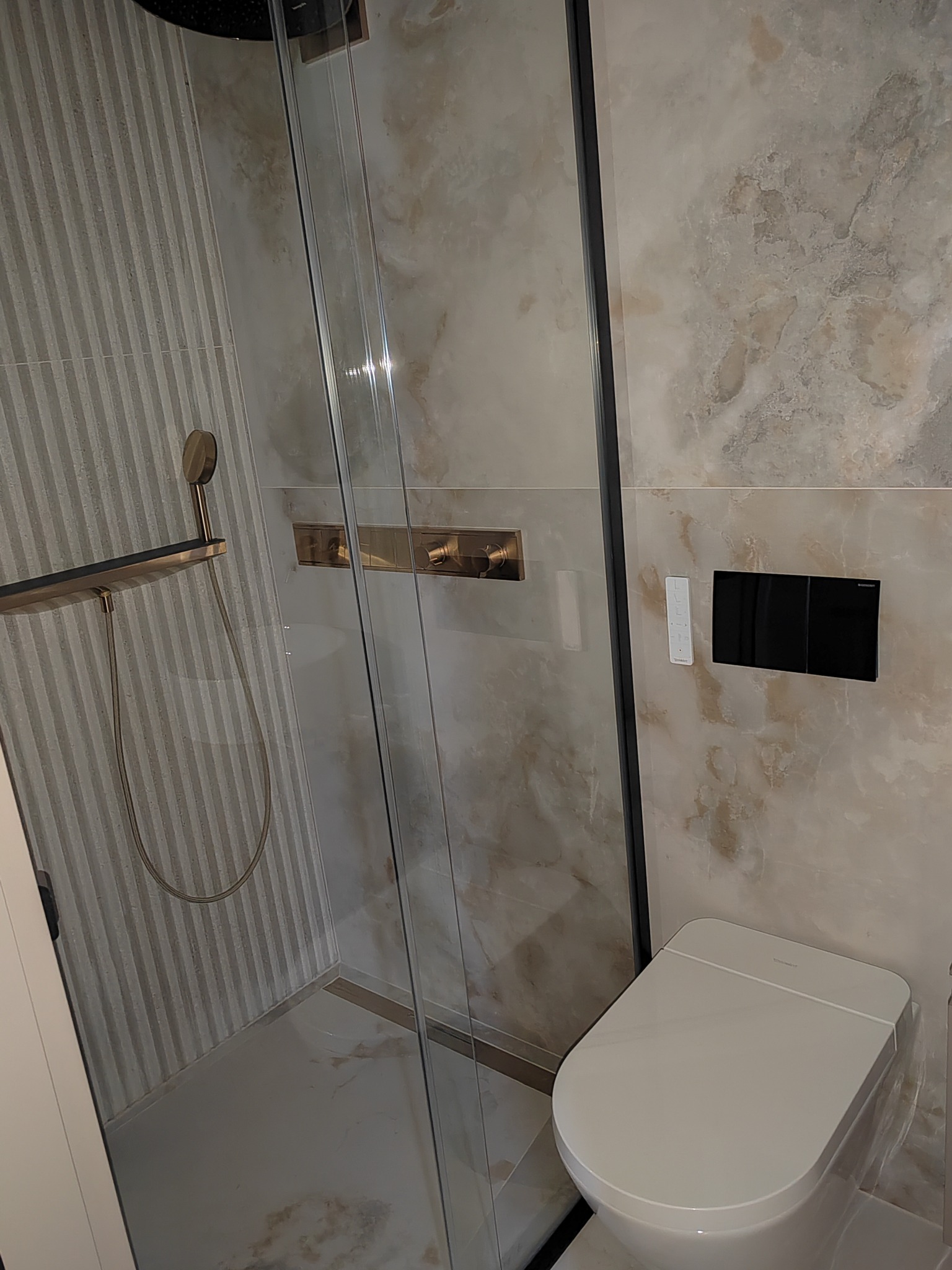 Nowoczesna łazienka z kabiną prysznicową typu walk-in, deszczownicą w kolorze szczotkowanego złota, podwieszaną miską WC i marmurowymi płytkami na ścianach.