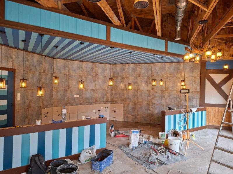 Wnętrze lokalu w trakcie wykończenia: drewniana konstrukcja sufitu, ściany z płytek, niebiesko-białe lady, wiszące lampy i narzędzia pracy na podłodze.