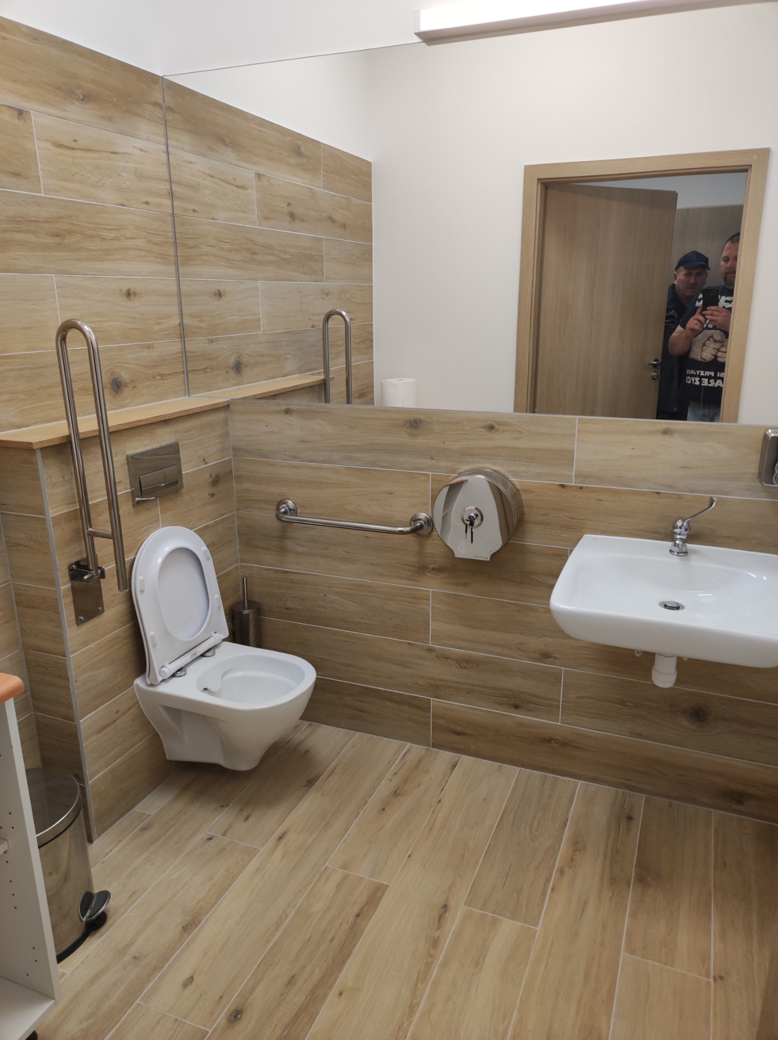 Jasna łazienka z drewnopodobnymi płytkami, toaleta z uchwytami dla niepełnosprawnych, umywalka i duże lustro odbijające fragment pomieszczenia z osobami.