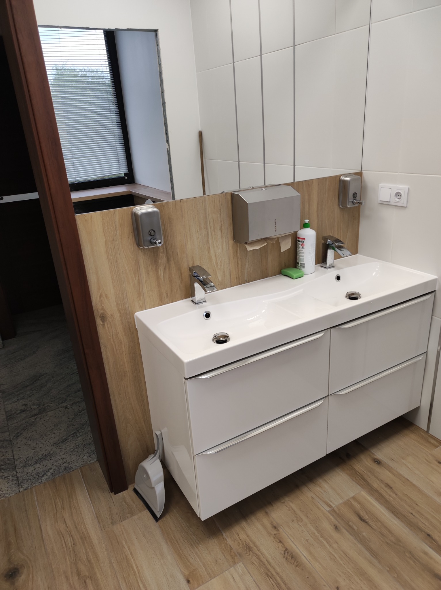 Nowoczesna łazienka z podwójną umywalką na szafce z szufladami, drewnianą podłogą i białymi płytkami. Widoczny dozownik mydła i okno z żaluzjami.