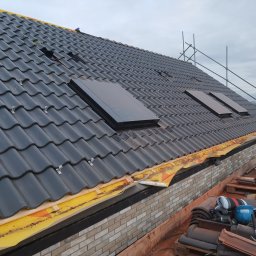 Iq Docieplenia Dachy - Czarny dach z dachówką karpiówką i trzema oknami dachowymi, widoczne zabezpieczenia żółtą folią na krawędzi dachu oraz rusztowanie w tle.