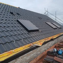 Czarny dach z dachówką karpiówką i trzema oknami dachowymi, widoczne zabezpieczenia żółtą folią na krawędzi dachu oraz rusztowanie w tle.