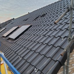 Iq Docieplenia Dachy - Czarny dach z dachówki ceramicznej w trakcie montażu, widoczne okna dachowe, rusztowanie z boku, oraz częściowo ułożone dachówki na górze dachu.