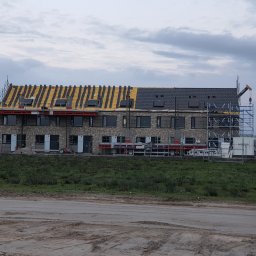 Iq Docieplenia Dachy - Dwukondygnacyjny budynek w trakcie budowy, z częściowo ukończonym dachem: jedna strona pokryta szarą dachówką, druga z widoczną żółtą membraną i łatami, rusztowania wzdłuż elewacji, teren budowy...