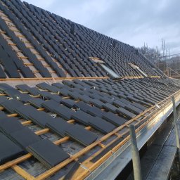 Iq Docieplenia Dachy - Układanie czarnej dachówki na łatach drewnianych, widoczne okno dachowe i elementy wentylacji, w tle rusztowanie i drzewa.