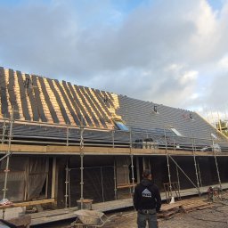 Iq Docieplenia Dachy - Częściowo pokryty dach z szarej dachówki, widoczne okna dachowe, rusztowania, pracownik w bluzie z logo firmy na dole kadru, przygotowania do dalszego krycia dachu.