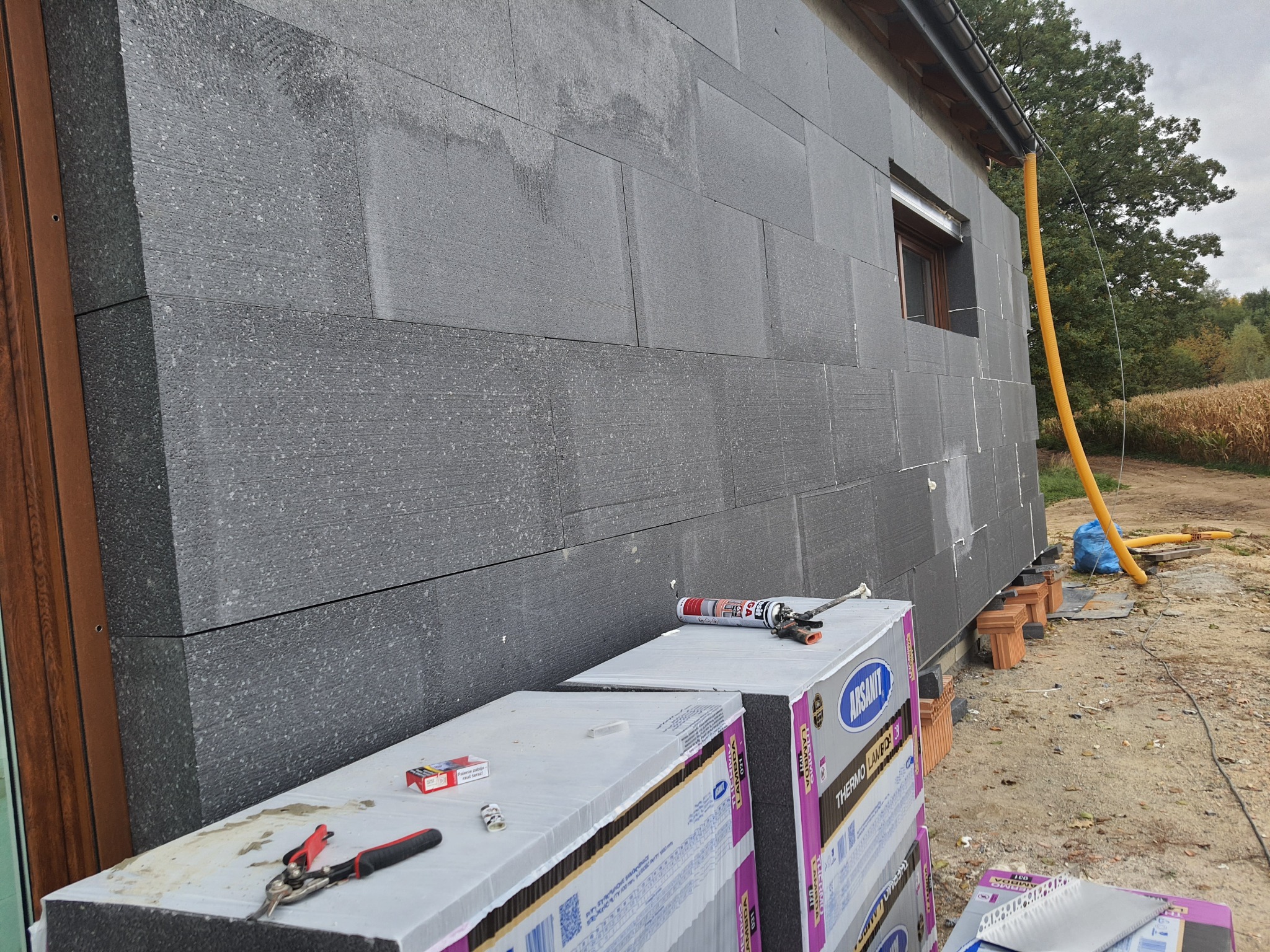 Ściana budynku w trakcie ocieplania szarym styropianem, widoczne puszki z pianką montażową, paczki styropianu ARSANIT Thermo Lamba 031, oraz narzędzia - kombinerki do cięcia blachy, żółta rura...