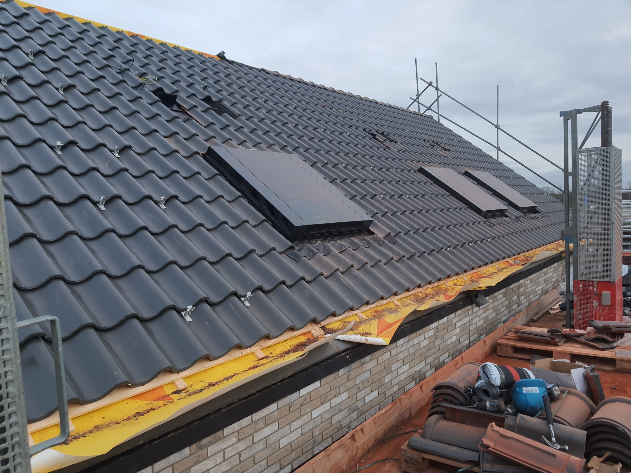 Czarny dach z dachówką karpiówką i trzema oknami dachowymi, widoczne zabezpieczenia żółtą folią na krawędzi dachu oraz rusztowanie w tle.