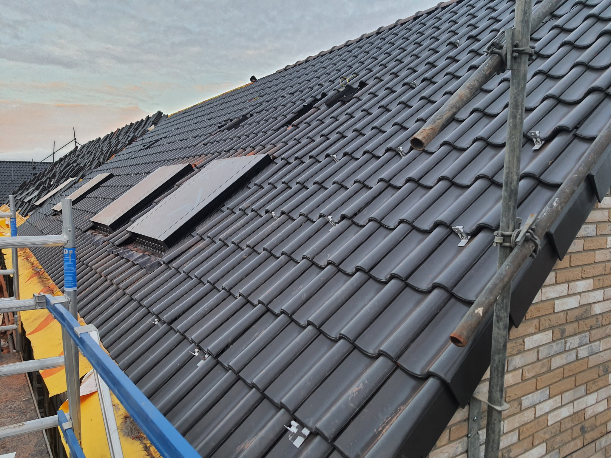 Czarny dach z dachówki ceramicznej w trakcie montażu, widoczne okna dachowe, rusztowanie z boku, oraz częściowo ułożone dachówki na górze dachu.