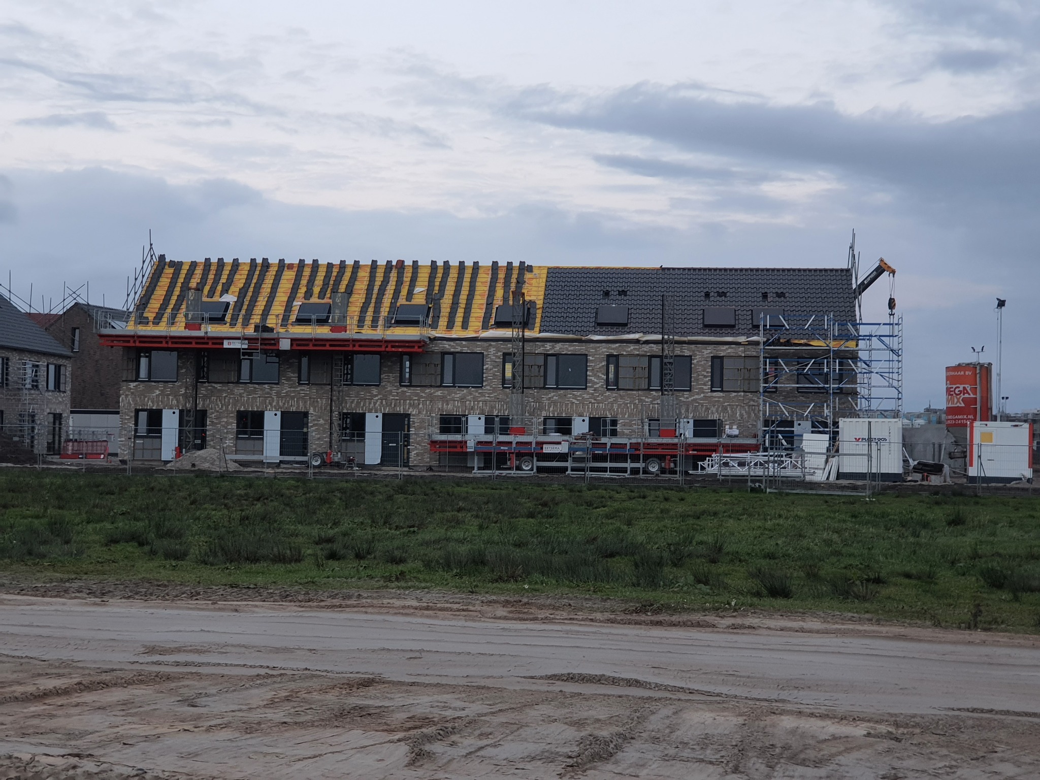 Dwukondygnacyjny budynek w trakcie budowy, z częściowo ukończonym dachem: jedna strona pokryta szarą dachówką, druga z widoczną żółtą membraną i łatami, rusztowania wzdłuż elewacji, teren budowy...