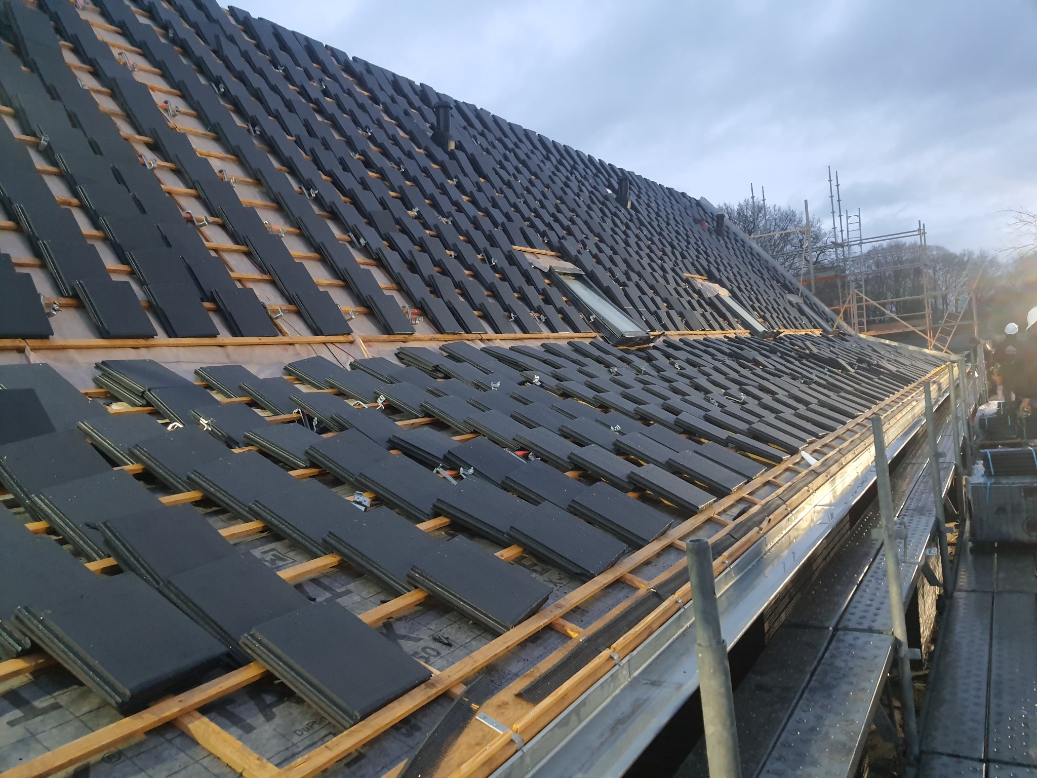 Układanie czarnej dachówki na łatach drewnianych, widoczne okno dachowe i elementy wentylacji, w tle rusztowanie i drzewa.