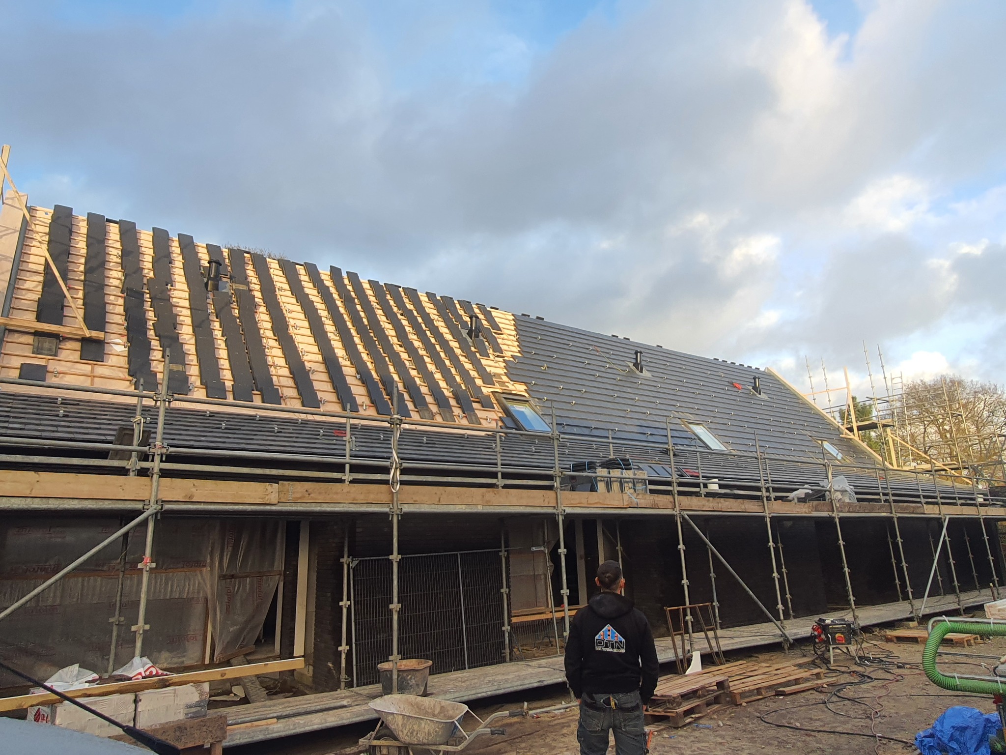 Częściowo pokryty dach z szarej dachówki, widoczne okna dachowe, rusztowania, pracownik w bluzie z logo firmy na dole kadru, przygotowania do dalszego krycia dachu.
