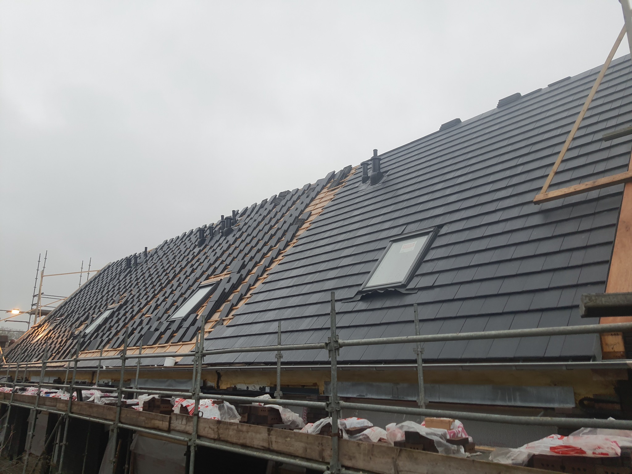 Montaż czarnej dachówki na skośnym dachu z oknami dachowymi, widoczne rusztowania i kominki wentylacyjne. Część dachu jeszcze niepokryta dachówką. Pochmurne niebo w tle.