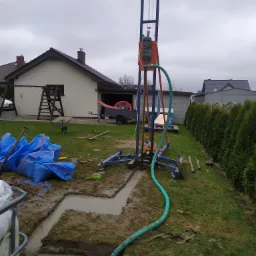 Wiercenie studni na posesji: widoczna wieża wiertnicza, wykopany rów z wodą, narzędzia i worki budowlane rozłożone na trawie, dom w tle, pochmurne niebo.