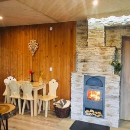 NOTO FLIZ - Drewniane wnętrze z kominkiem obłożonym kamieniem, jasne meble, dekoracje na ścianie, płomienie w palenisku, drewno ułożone pod kominkiem, bukiet kwiatów na kominku.