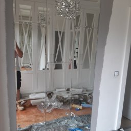 NOTO FLIZ - Montaż lustrzanych paneli na ścianie w pokoju z białymi szafami wnękowymi, widoczne narzędzia i materiały budowlane na podłodze, częściowo zakrytej folią ochronną.