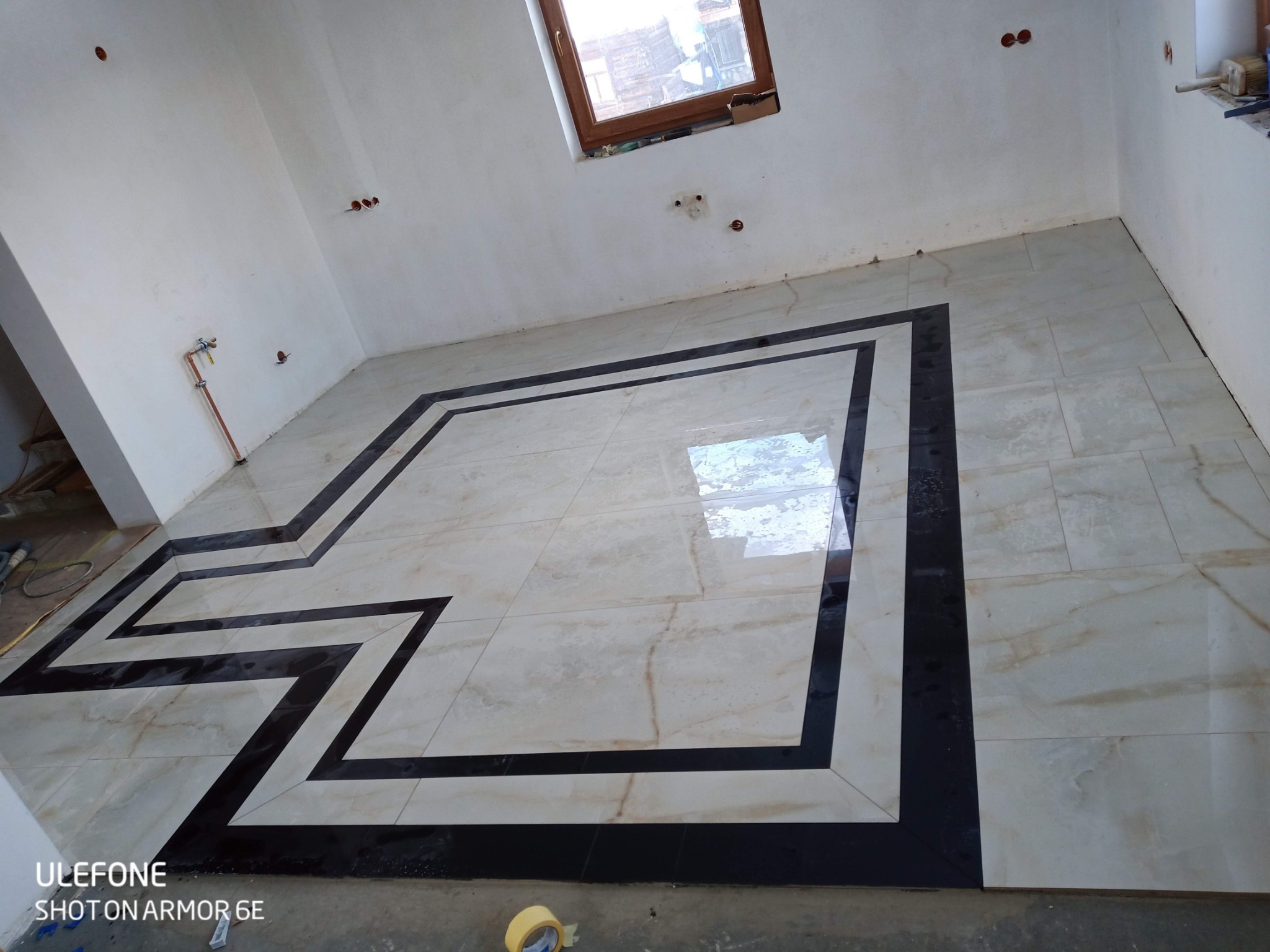 Ułożona podłoga z jasnych płytek z czarną bordiurą w kształcie zygzaka, widoczne niedokończone ściany i instalacje elektryczne.