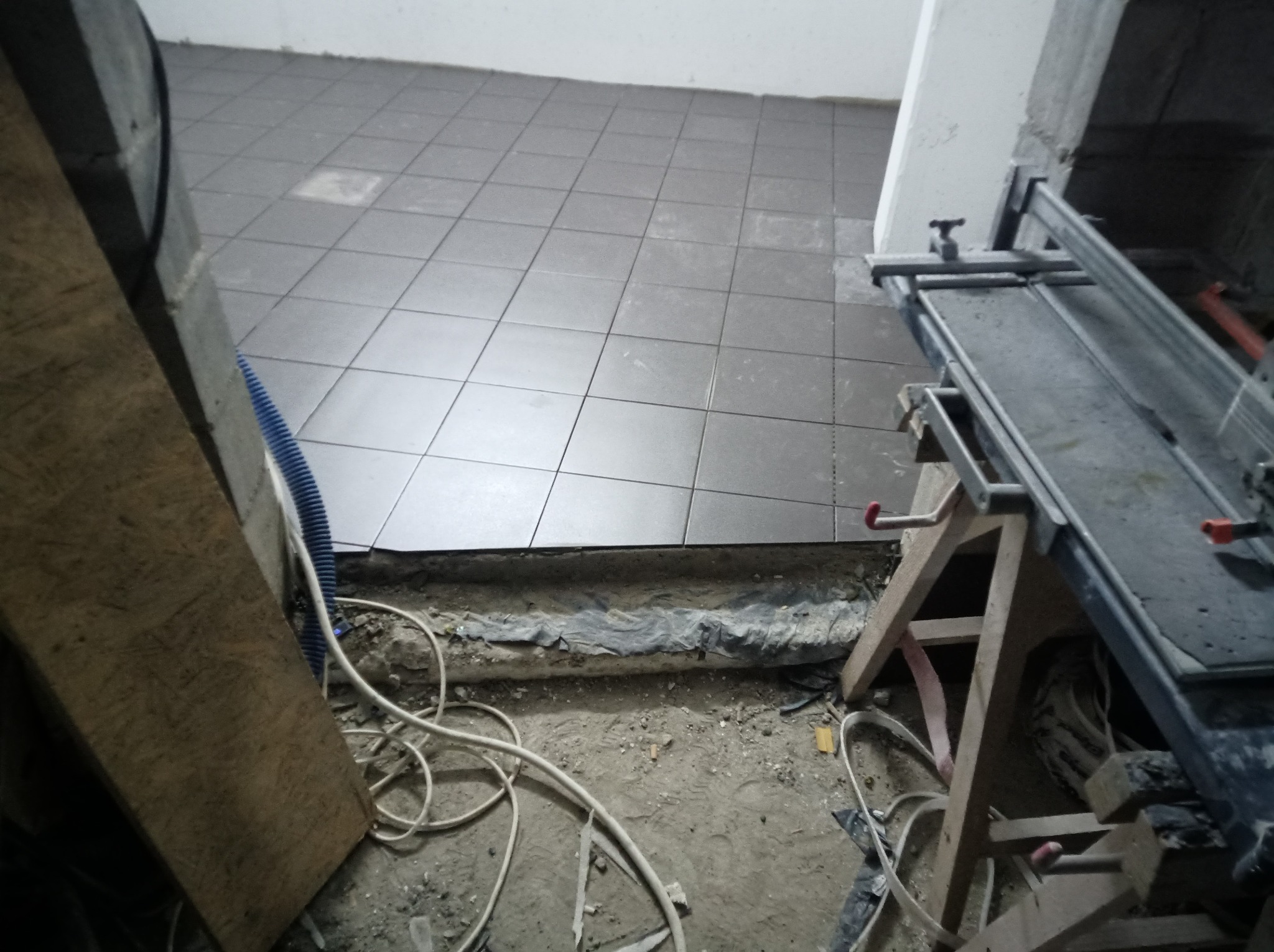 Proces układania szarych płytek podłogowych w pomieszczeniu, widoczna krawędź ułożonej podłogi, przygotowane podłoże z folią, białe kable i narzędzie do cięcia płytek.