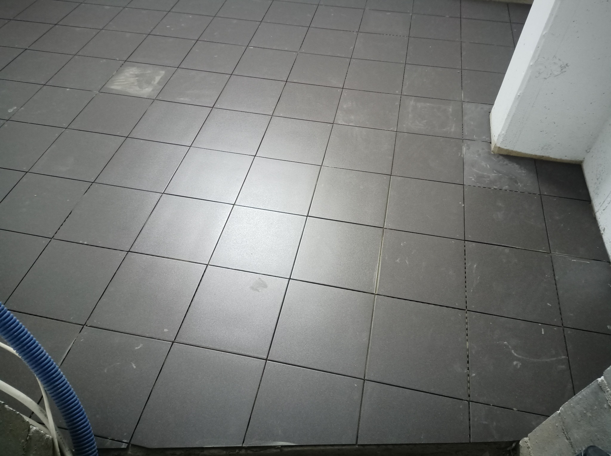 Ułożone kwadratowe, grafitowe płytki podłogowe z widocznymi fugami, fragment kolumny i niebieski wąż odkurzacza w rogu.