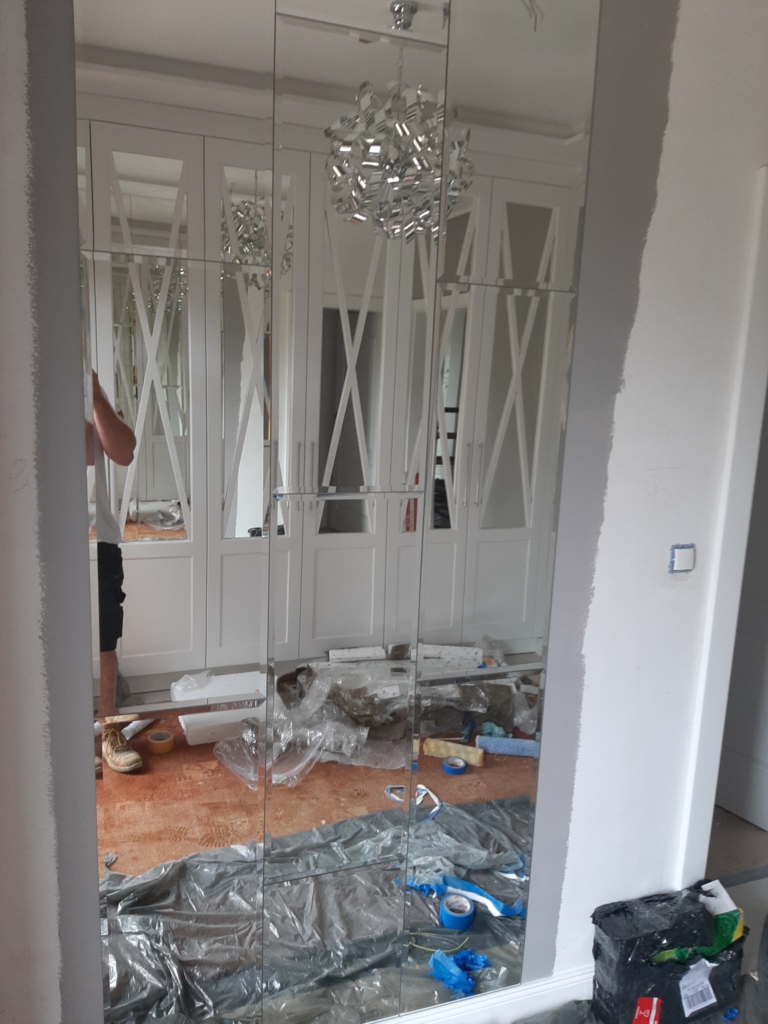 Montaż lustrzanych paneli na ścianie w pokoju z białymi szafami wnękowymi, widoczne narzędzia i materiały budowlane na podłodze, częściowo zakrytej folią ochronną.
