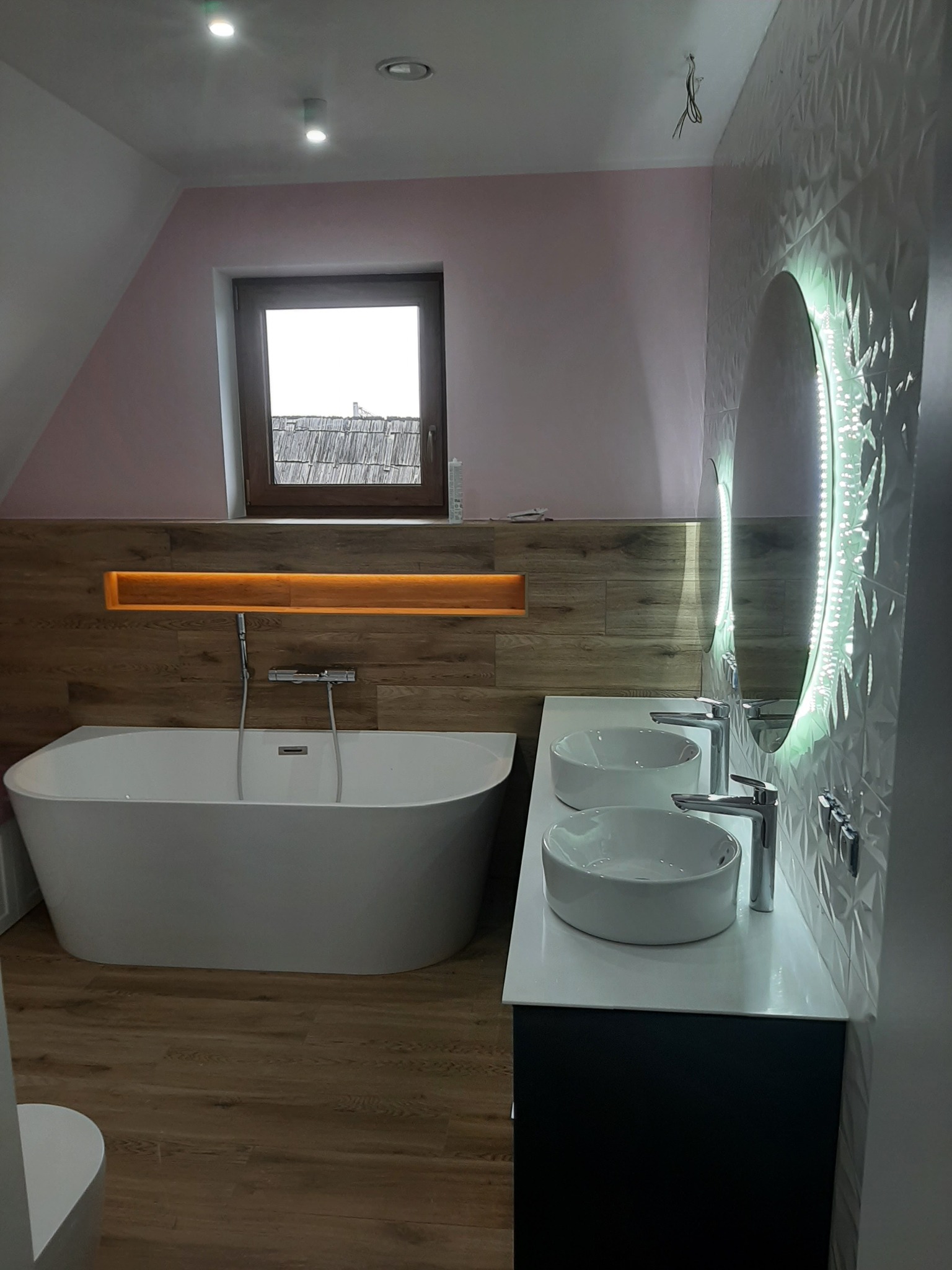 Nowoczesna łazienka na poddaszu z wolnostojącą wanną, dwiema umywalkami nablatowymi, podświetlanym lustrem i drewnopodobnymi płytkami na podłodze oraz ścianie w strefie wanny.