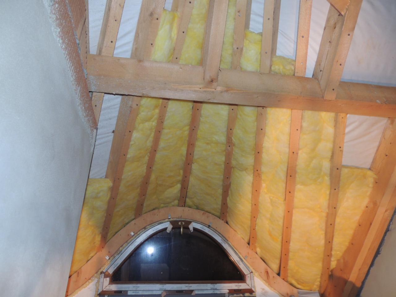 Izolacja poddasza wełną mineralną między krokwiami, widoczne okno dachowe w kształcie łuku i fragment ściany pokrytej pianą PUR.