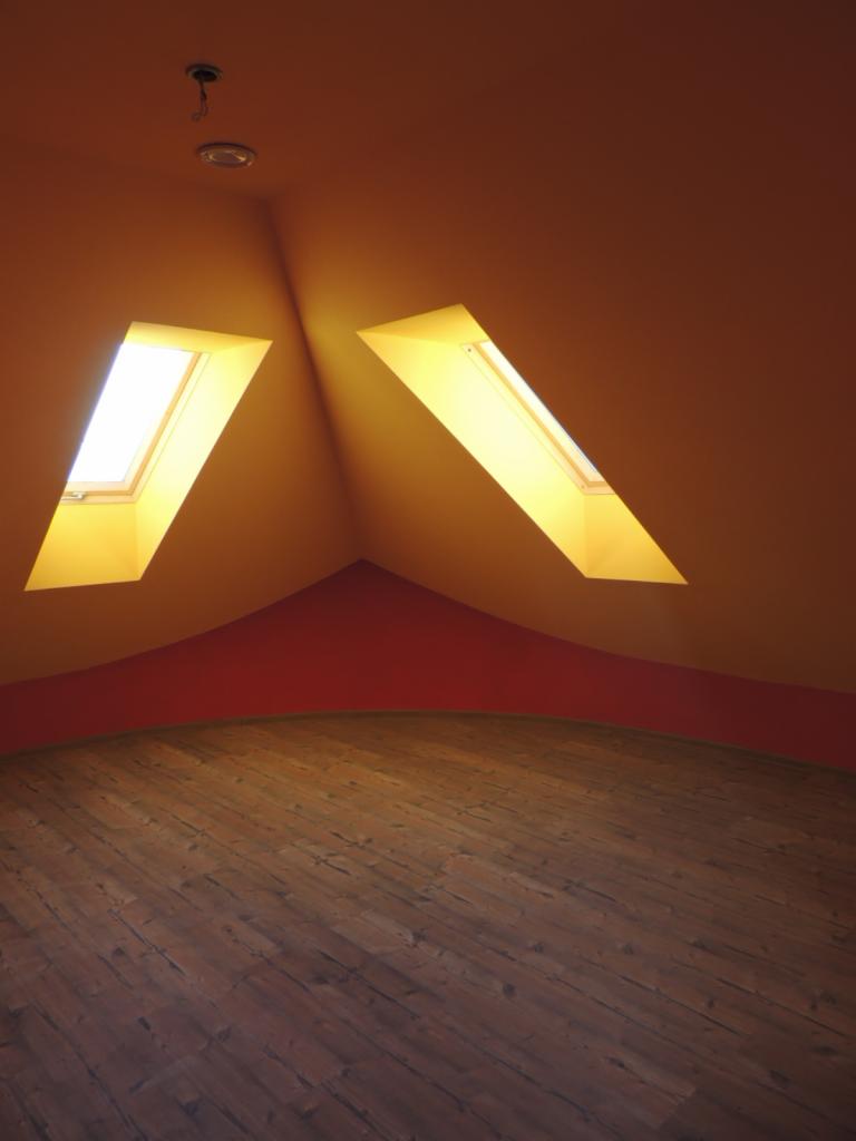 Poddasze z dwoma oknami dachowymi, pomalowane na pomarańczowo z czerwonym pasem przy podłodze, drewniana podłoga z desek, widok na surowy montaż oświetlenia.