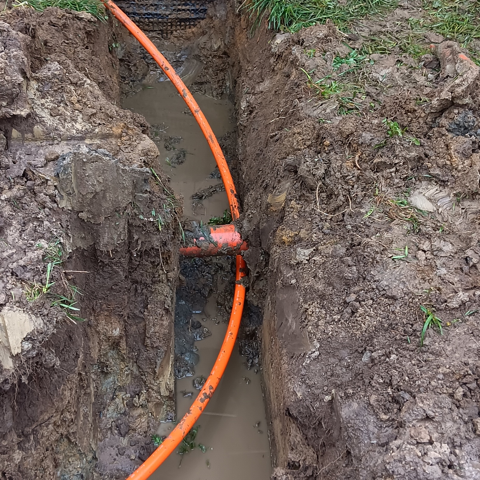 Pomarańczowy przewód w ziemnym wykopie wypełnionym wodą, widoczne połączenie rur.
