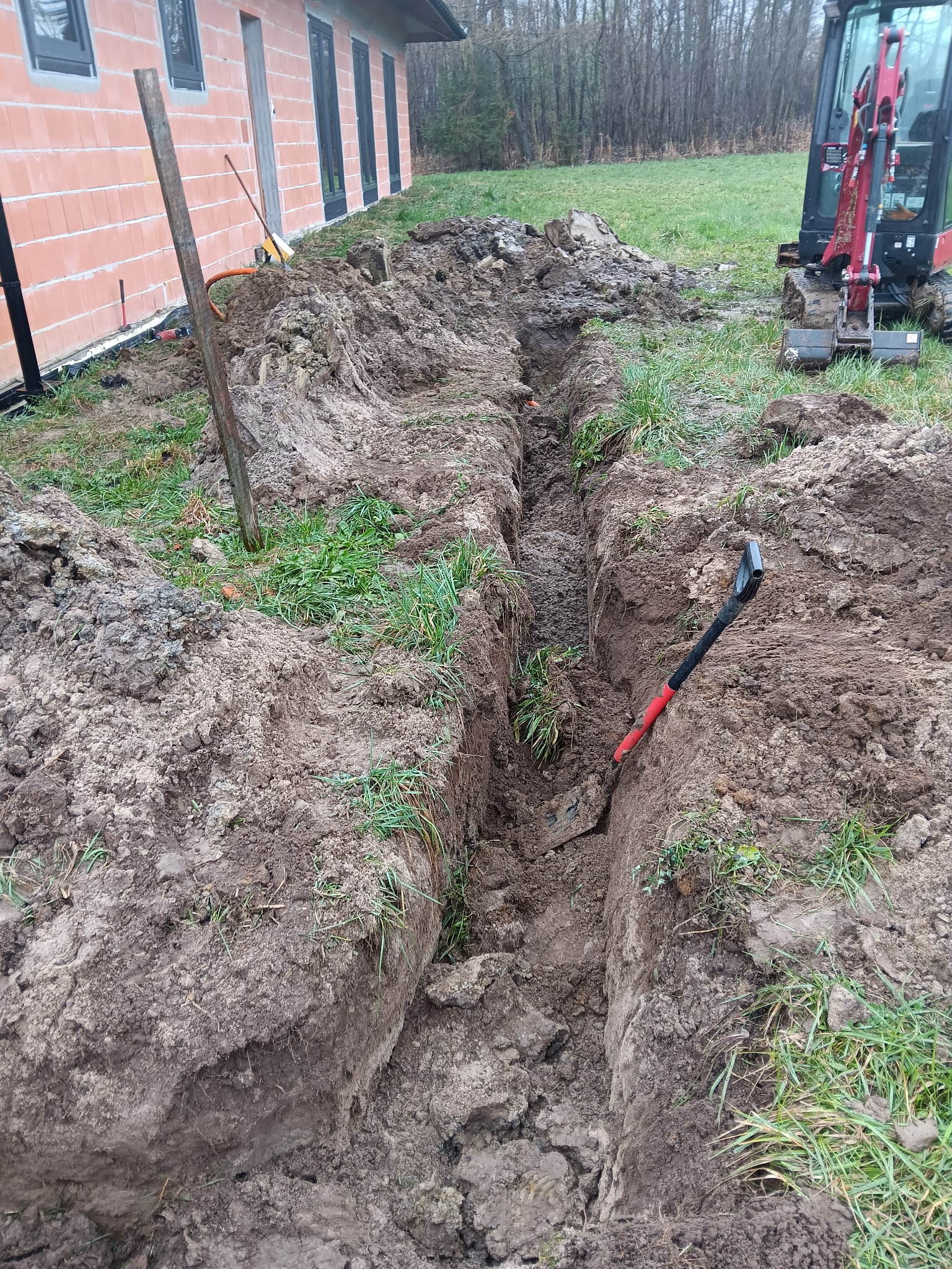 Wykopany wąski rów w ziemi wzdłuż ściany budynku z czerwonej cegły, z mini koparką i łopatą w tle.