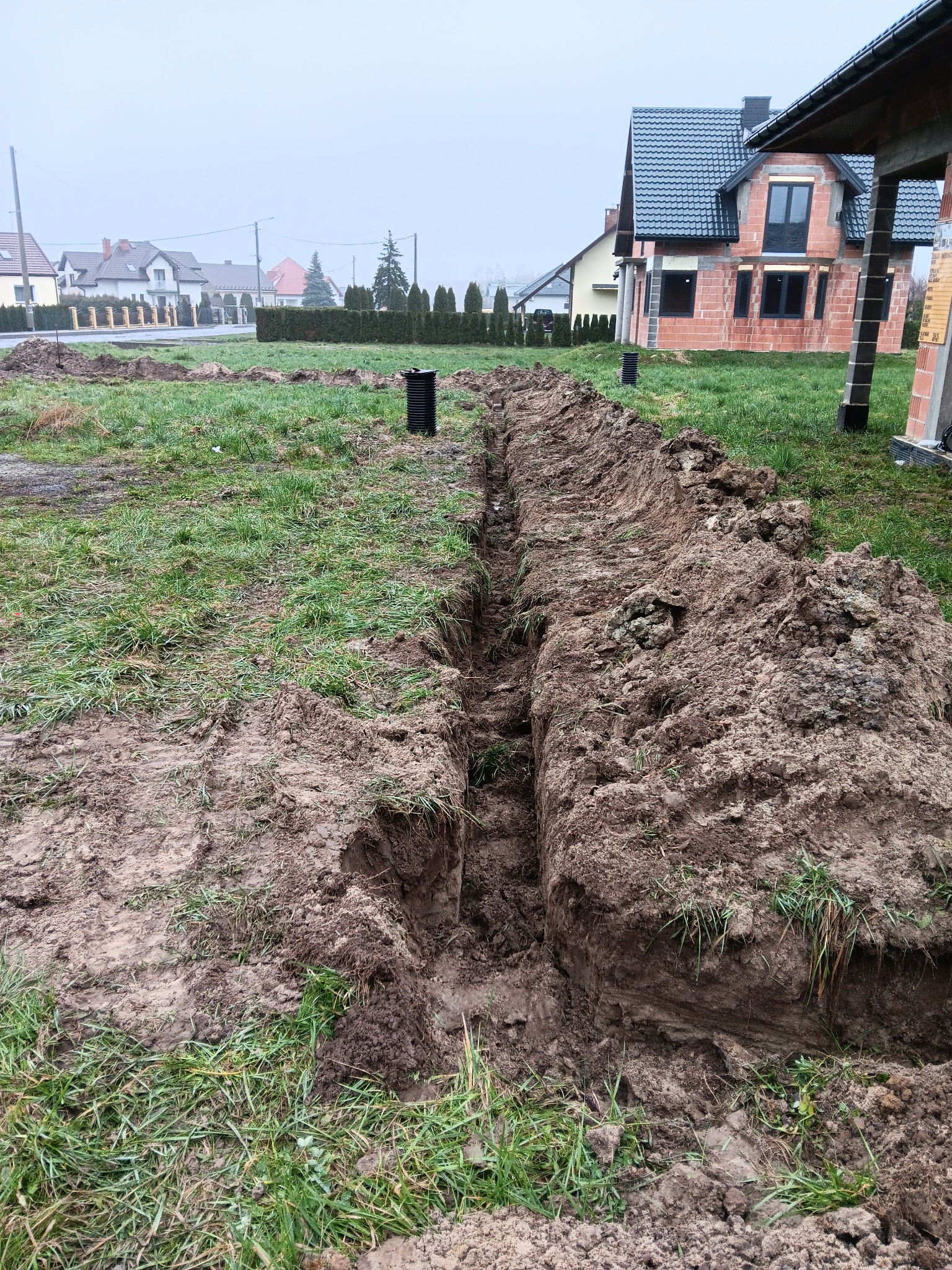 Długi, wąski wykop w ziemi na działce budowlanej, widoczny czarny element kanalizacji, w tle dom w trakcie budowy z czerwonej cegły i zabudowa jednorodzinna w oddali, pochmurny dzień.