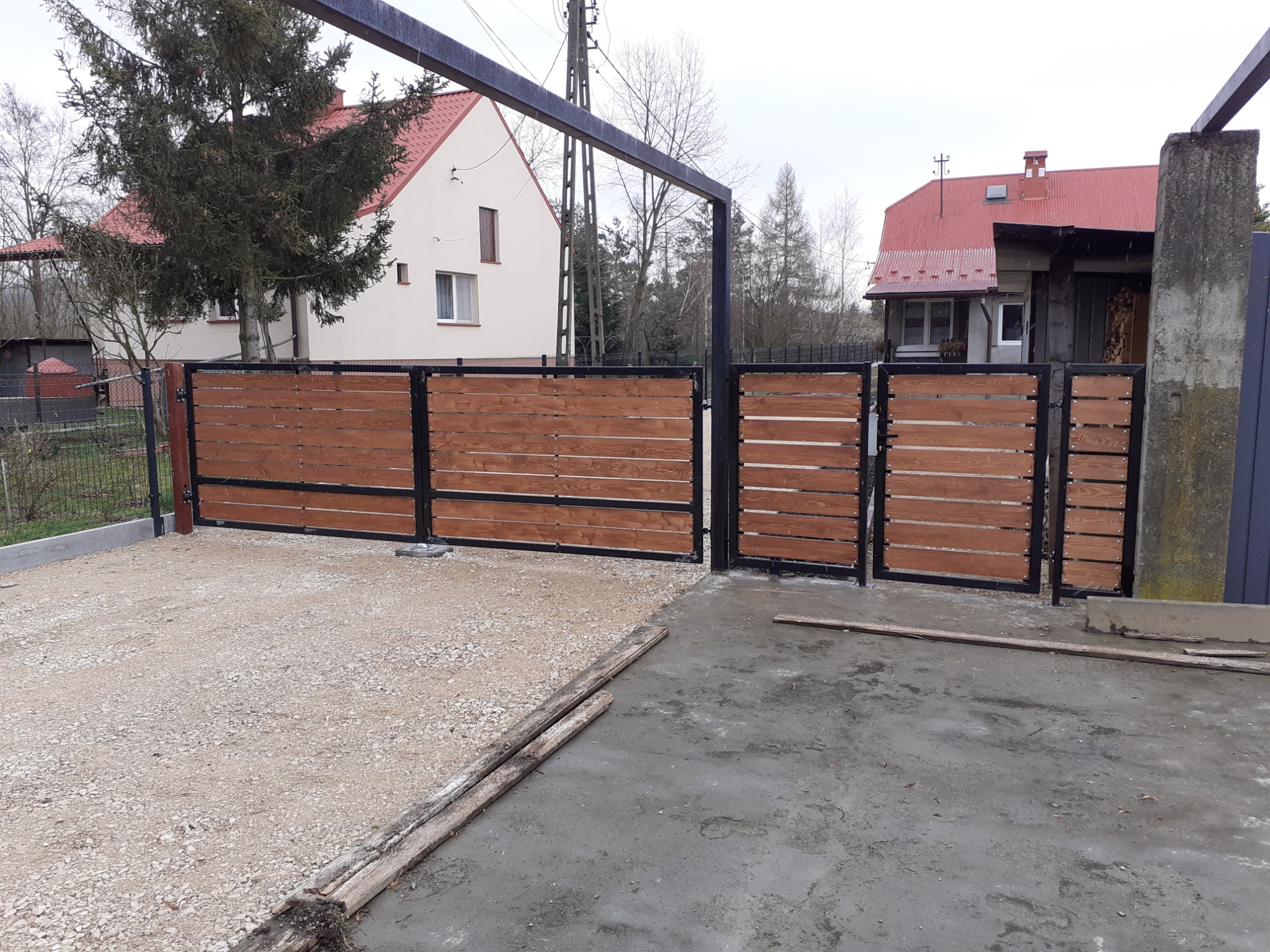 Nowoczesna brama wjazdowa i furtka z drewnianymi panelami w kolorze ciepłego brązu i czarną metalową ramą, na tle dwóch domów o różnej architekturze i szarego nieba.