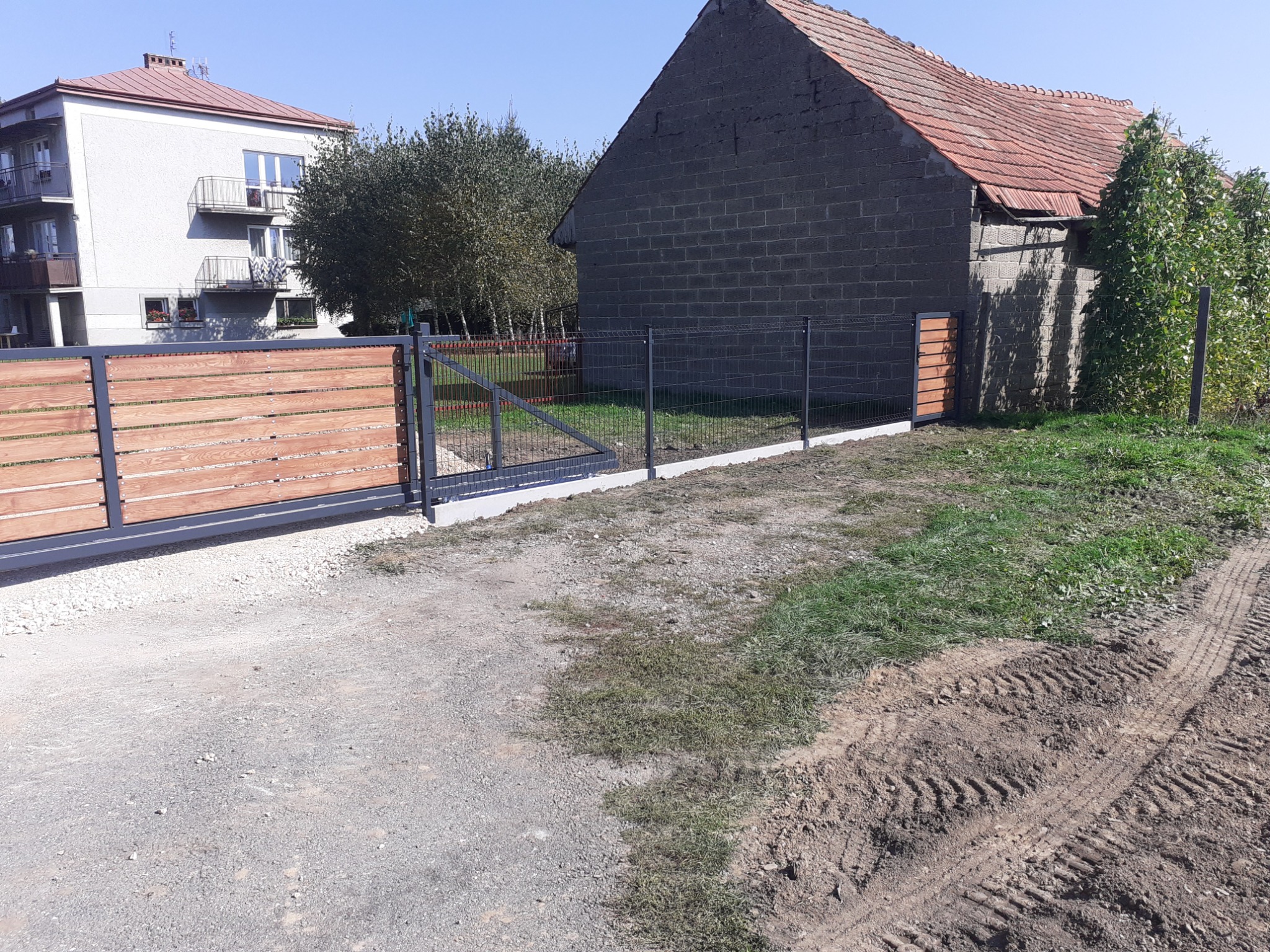Nowoczesna brama przesuwna z drewnianymi panelami i metalowa siatka ogrodzeniowa na tle budynku mieszkalnego i starej stodoły z ubytkami w dachu, widok z drogi gruntowej z wyraźnymi śladami opon.