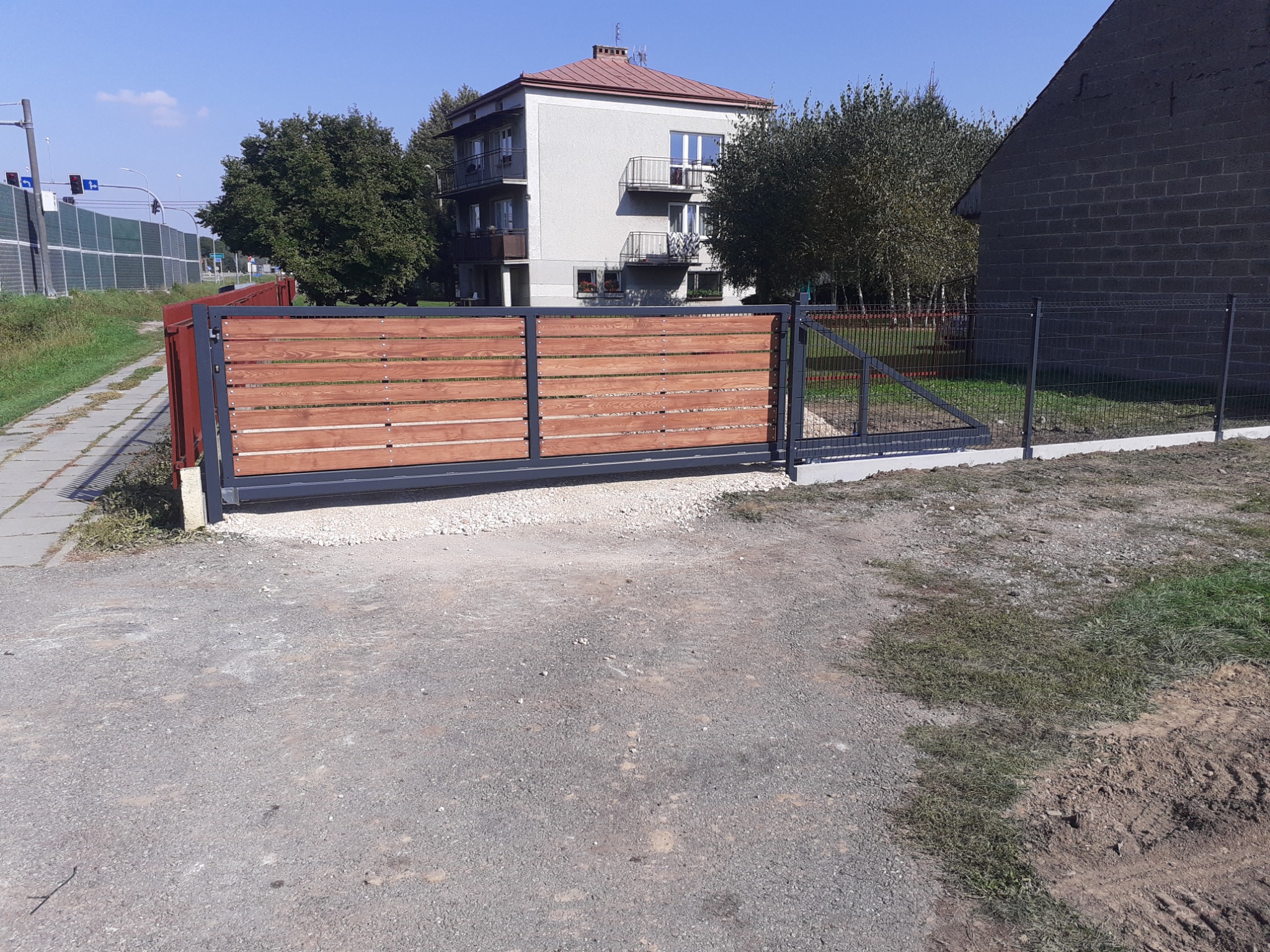 Nowoczesna, przesuwna brama wjazdowa z drewnianymi panelami i furtką, otoczona siatką ogrodzeniową na betonowej podmurówce, z budynkiem mieszkalnym w tle i widoczną sygnalizacją świetlną.