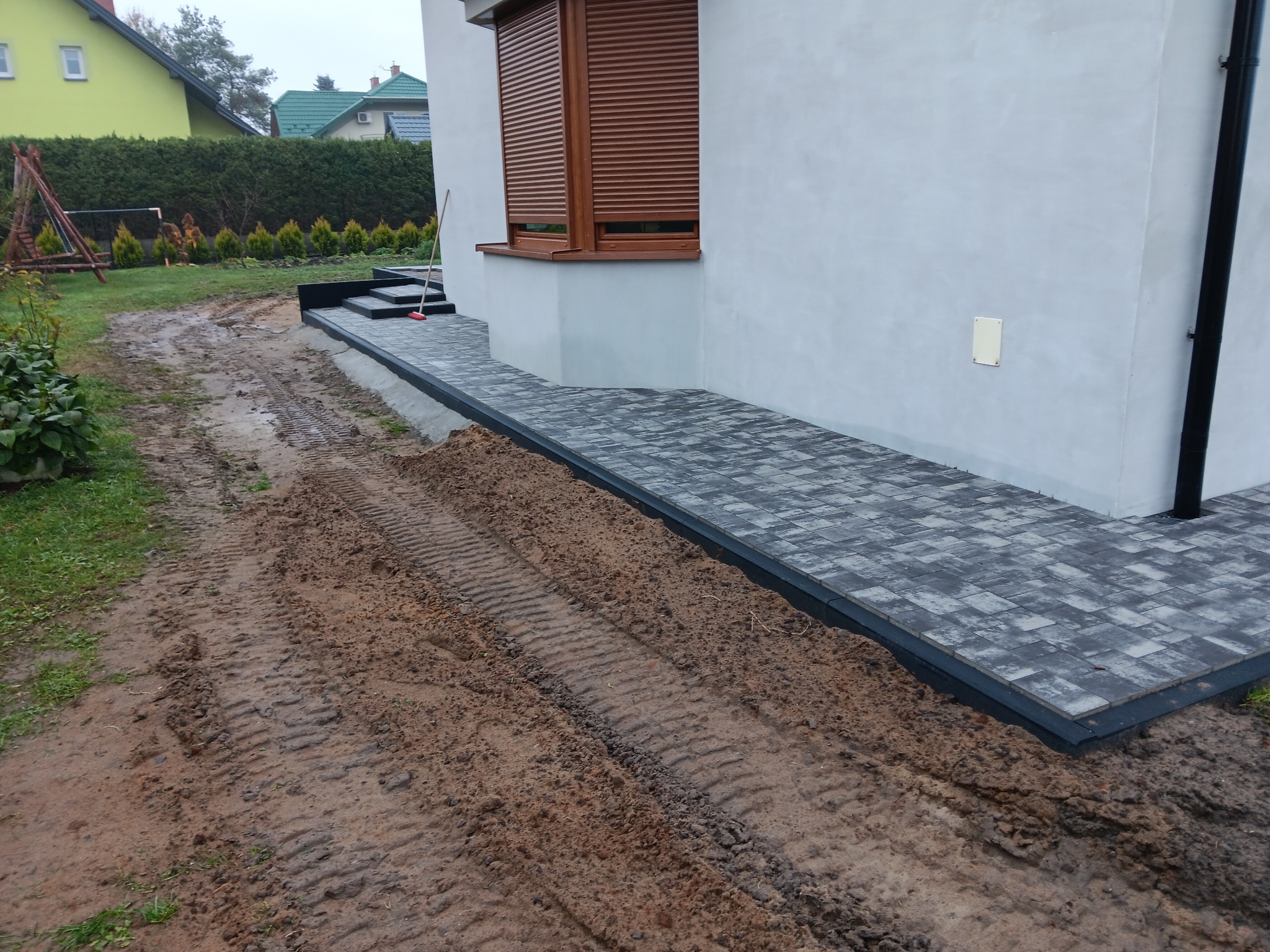 Ułożona kostka brukowa przy ścianie domu z brązowymi roletami, widoczne ślady opon na ziemi i trawnik w tle.