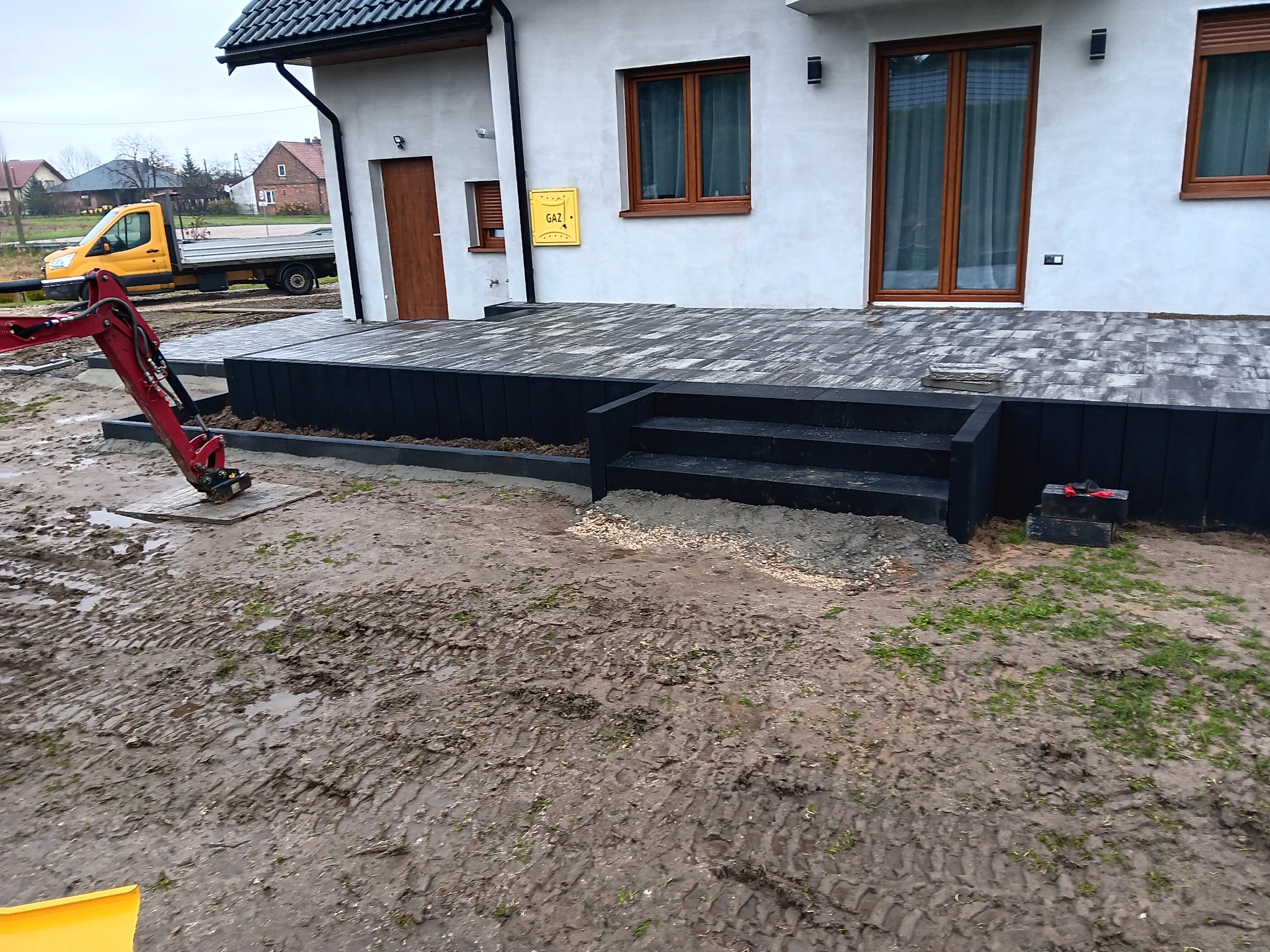 Mini koparka przy układaniu kostki brukowej na tarasie przed domem z czarnymi schodami, widoczny żółty samochód dostawczy w tle.