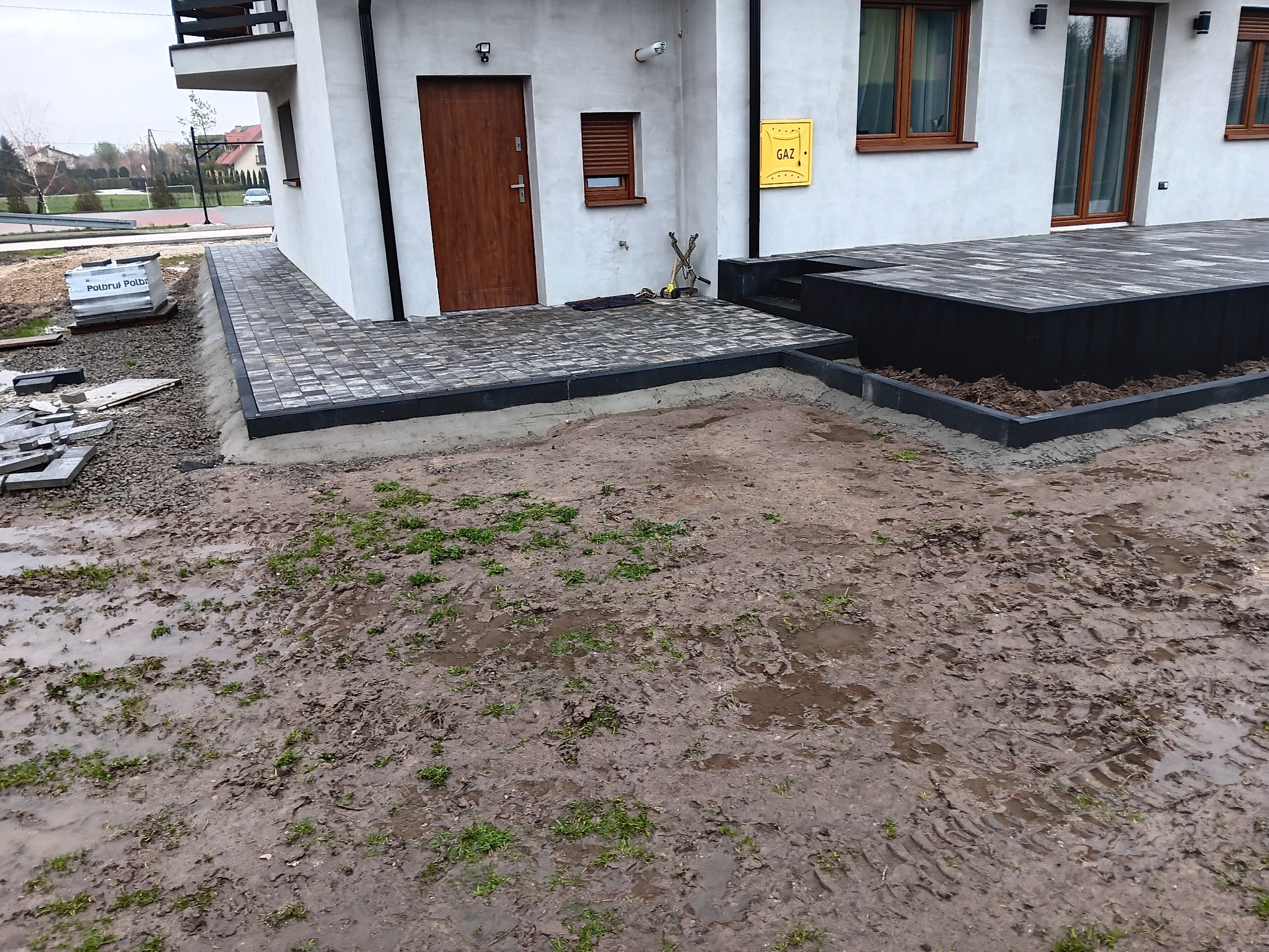 Układanie kostki brukowej przed domem jednorodzinnym, widoczny fragment chodnika z kostki, taras z kostki na podwyższeniu, błotniste podłoże przed domem, paleta z kostką brukową w tle.