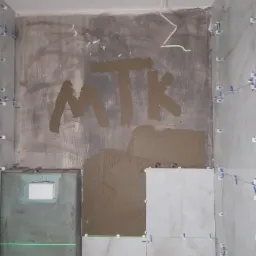 Wnętrze łazienki w trakcie układania płytek, widoczna ściana z napisem 'MTK' wykonanym z zaprawy, fragment ściany z nałożonym klejem do płytek, laserowy poziom wyznaczający linię, białe płytki...