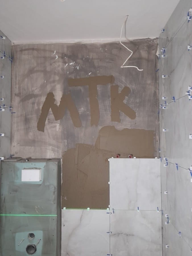 Wnętrze łazienki w trakcie układania płytek, widoczna ściana z napisem 'MTK' wykonanym z zaprawy, fragment ściany z nałożonym klejem do płytek, laserowy poziom wyznaczający linię, białe płytki...
