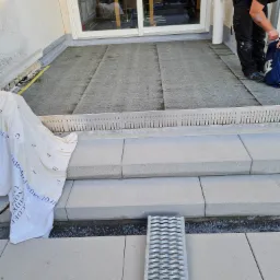 Montaż schodów zewnętrznych z jasnoszarych płyt kamiennych, z widocznym systemem odwodnienia liniowego i pracownikiem w tle, na tarasie z papy.