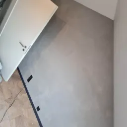 Ściana pokryta szarym tynkiem dekoracyjnym, widoczne gniazdka elektryczne, fragment podłogi z parkietem w jodełkę i białe drzwi z metalową klamką.