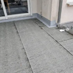 Szare, bitumiczne płyty na płaskim dachu, widoczne połączenia, obróbka blacharska, rynna z filtrem, konewka i wiadro w tle.