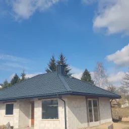 Nowy, grafitowy dach z blachodachówki na parterowym domu z białego bloczka, widok z dołu na tle błękitnego nieba z obłokami.