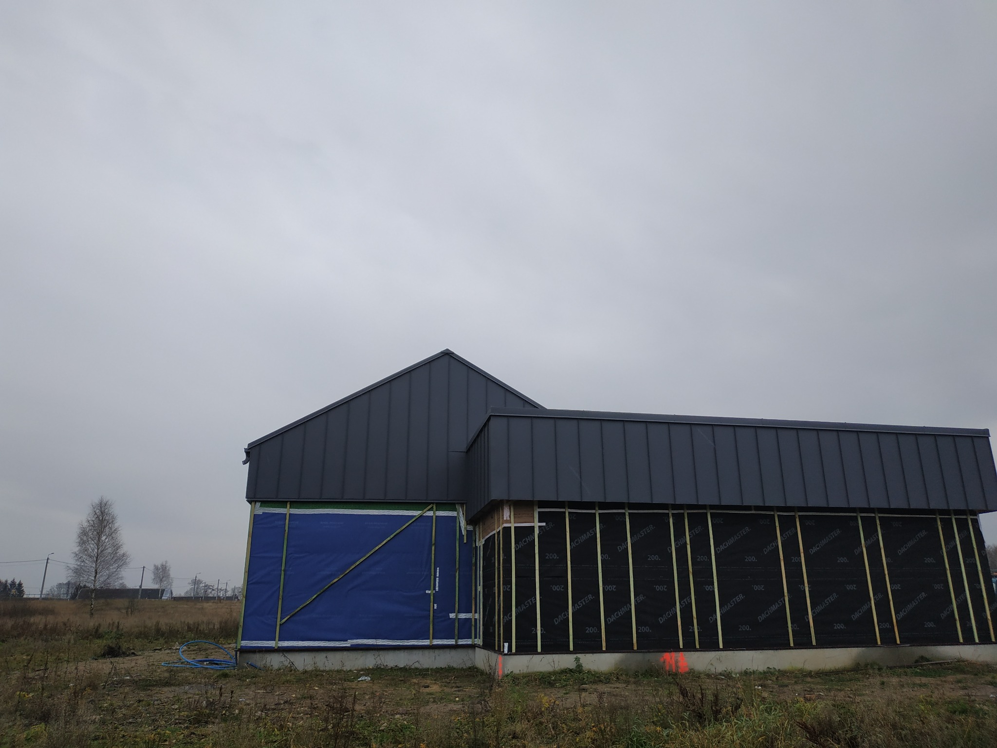 Budynek w trakcie budowy, z widocznym fragmentem dachu z blachy na rąbek stojący i przygotowaną ścianą z membraną Dachmaster oraz listwami drewnianymi, teren wokół niezagospodarowany.