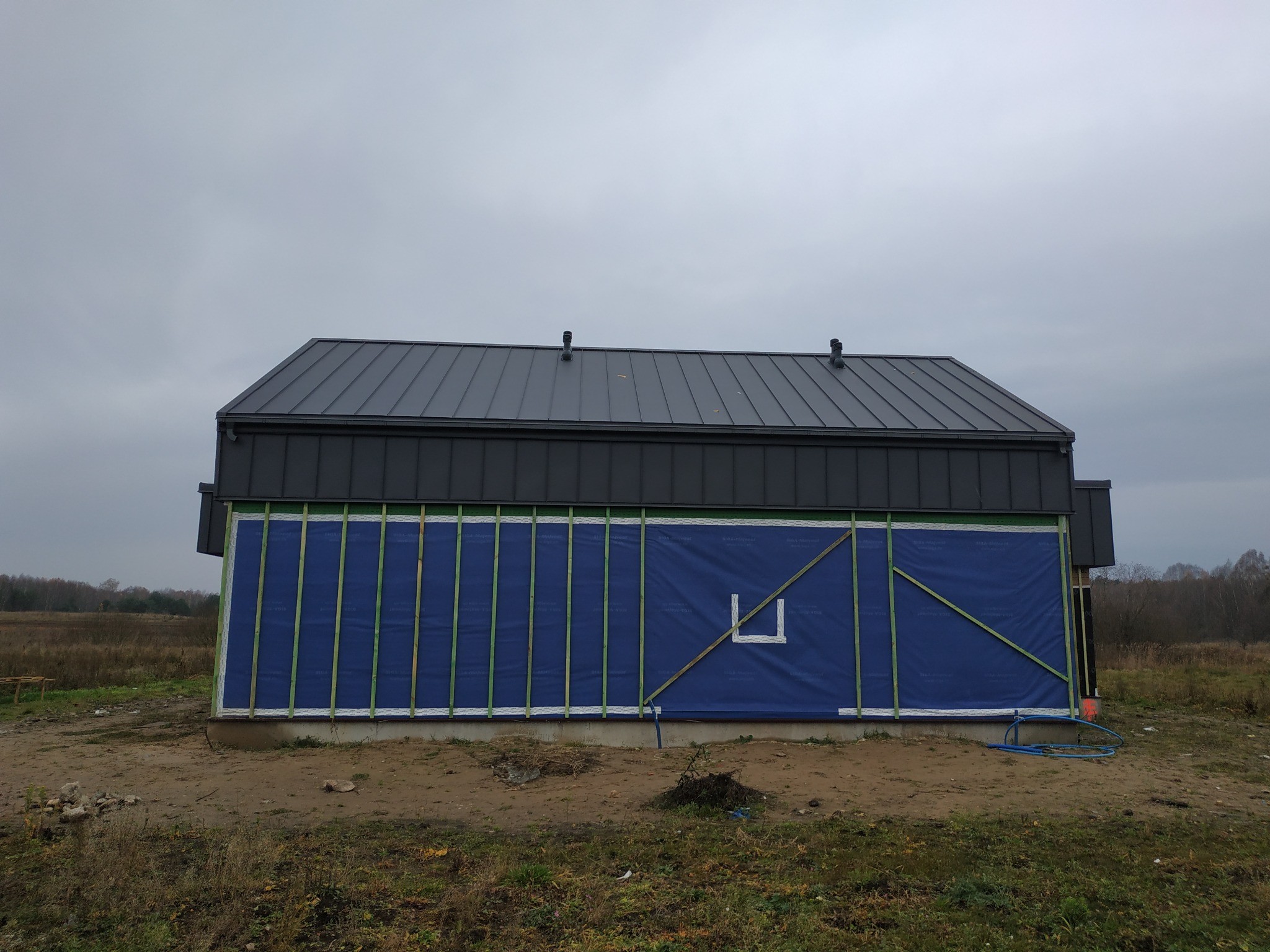 Budynek w trakcie budowy z zamontowanym ciemnoszarym dachem z blachy na rąbek stojący, zabezpieczony niebieską membraną wiatroizolacyjną i zieloną konstrukcją drewnianą pod elewację, widoczne...