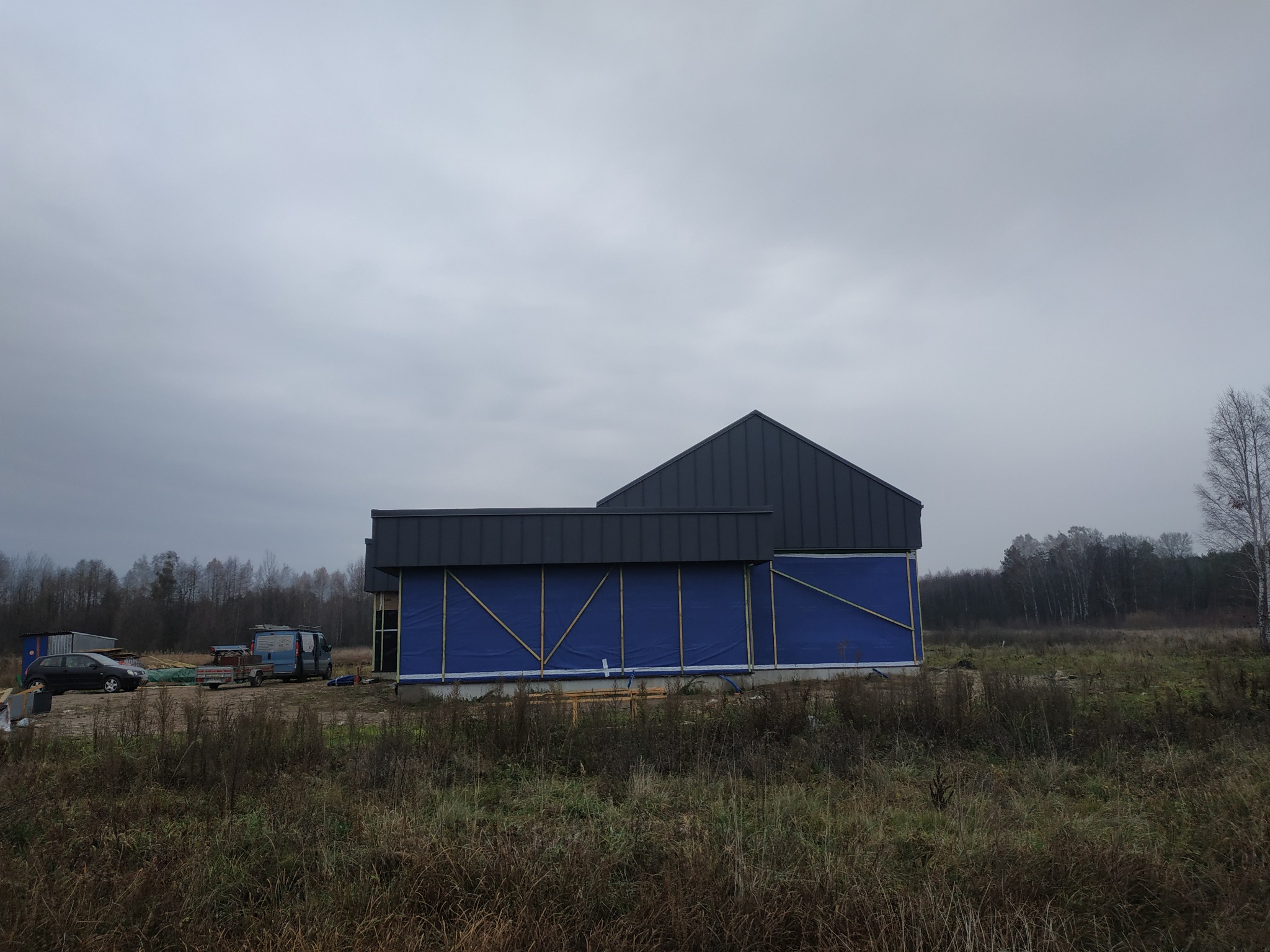 Budynek w trakcie budowy z zamontowanym dachem z blachy na tle pochmurnego nieba, ściany zabezpieczone niebieską folią i drewnianymi łatami, widoczne otoczenie budowy z pojazdami i drzewami.