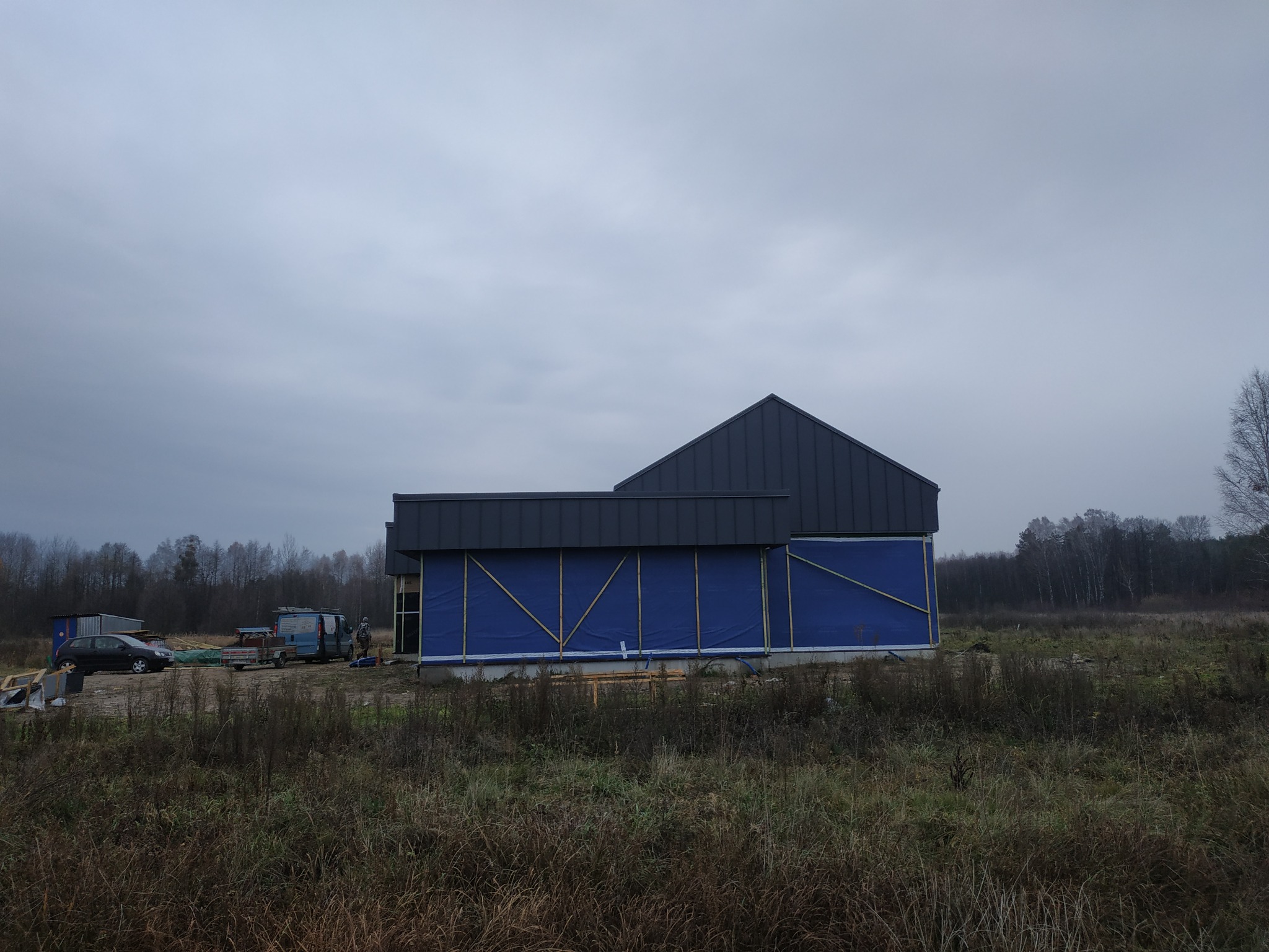 Dom w trakcie budowy, z widoczną konstrukcją dachu i niebieskim zabezpieczeniem ścian, na tle pochmurnego nieba i jesiennego krajobrazu.