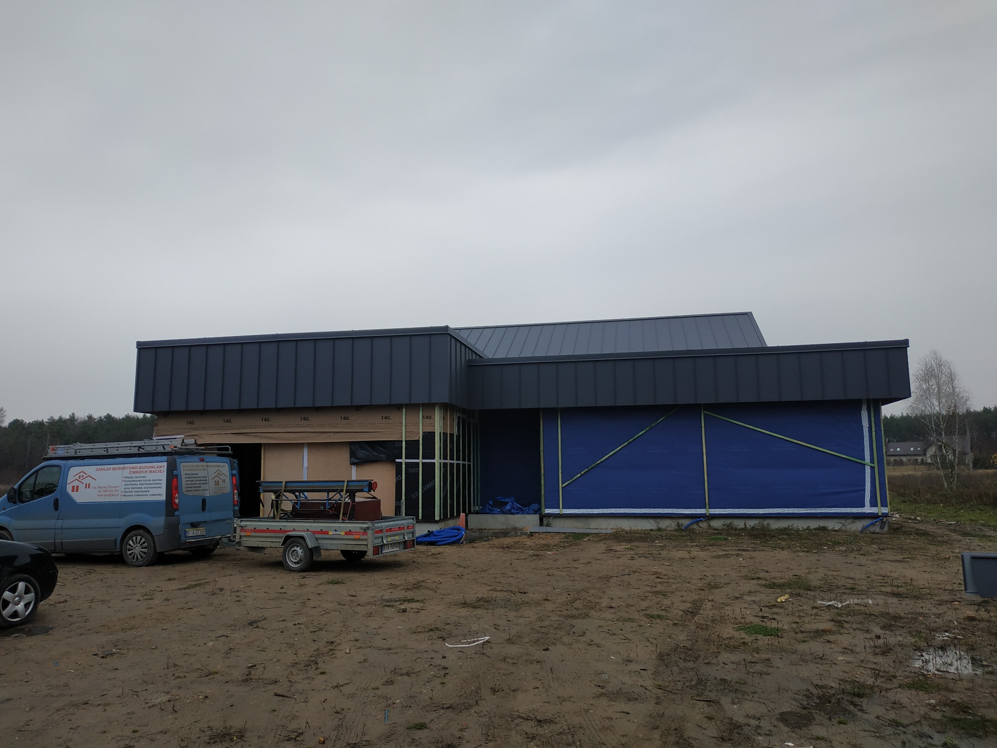 Budynek w trakcie budowy z nowym, grafitowym dachem, zabezpieczony folią budowlaną, na placu budowy zaparkowany samochód dostawczy z reklamą firmy dekarskiej i przyczepka.