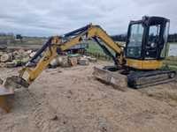 Żółta minikoparka na piaszczystym podłożu, w tle stosy kostki brukowej i drzewa pod pochmurnym niebem.