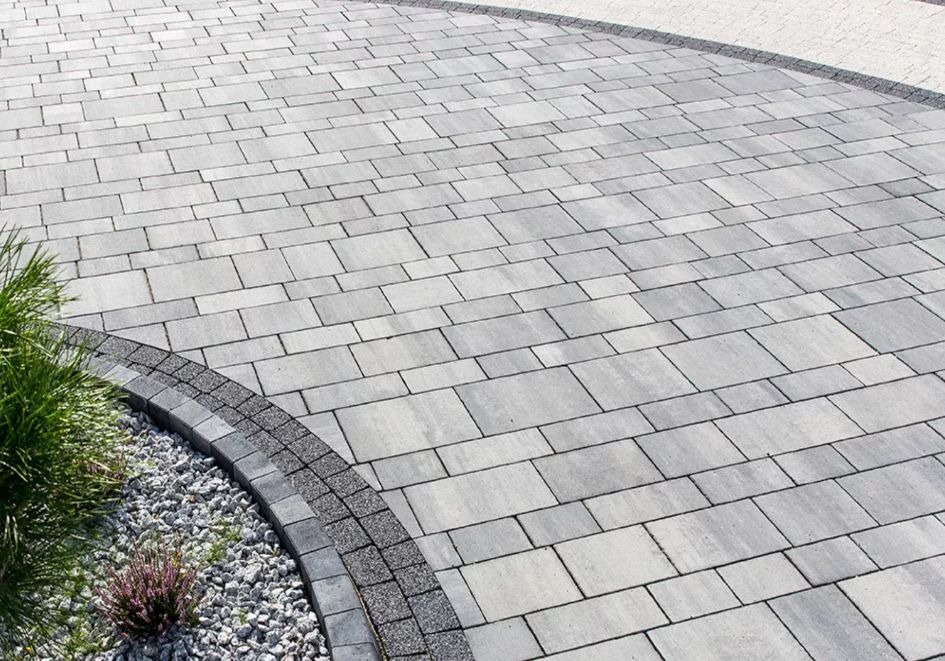 Ułożona szara kostka brukowa w kształcie prostokątów, obrzeże z ciemniejszych kostek i jasnych kamyków, trawa ozdobna i mała roślina z fioletowymi kwiatami.