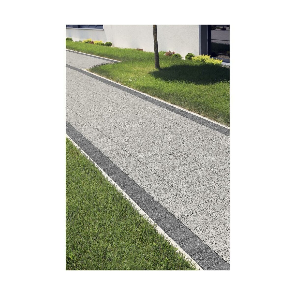 Szeroki, szary chodnik z kostki brukowej z ciemnoszarym obramowaniem, otoczony zielonym trawnikiem i elementami architektury ogrodowej.